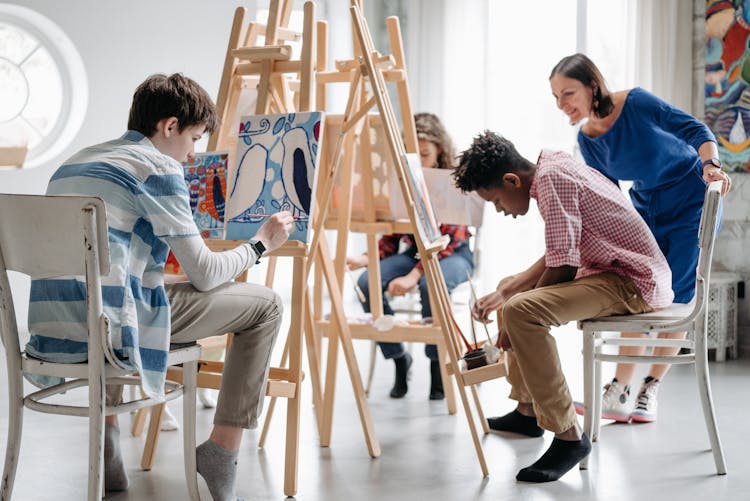 A group of young students painting on easels with a teacher encouraging them.