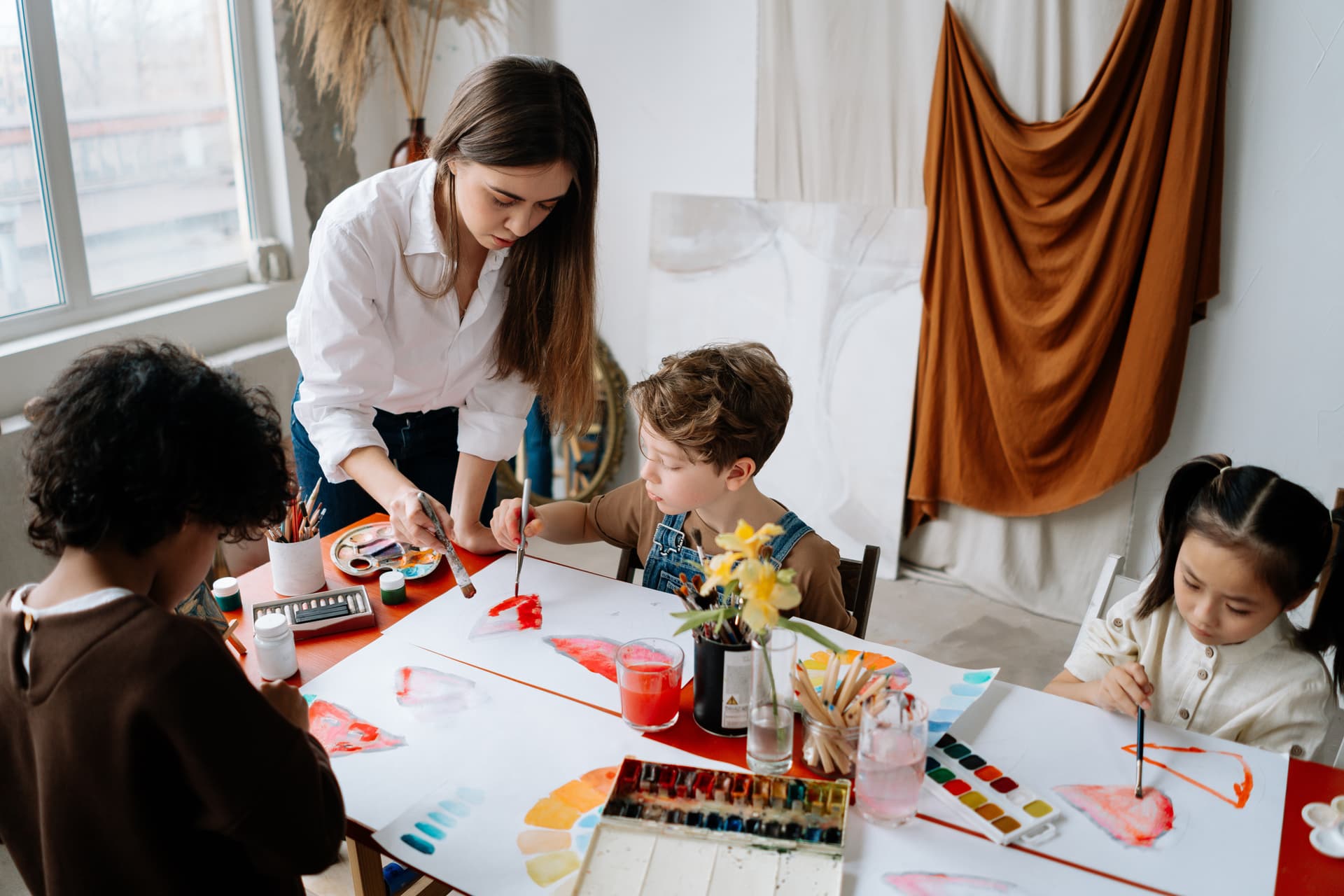 Teacher helping children with art supplies. Children are painting on white paper.