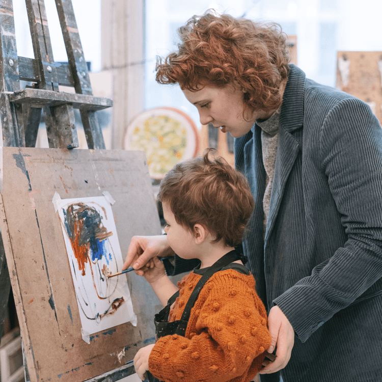 Woman helping young child paint on an easel.