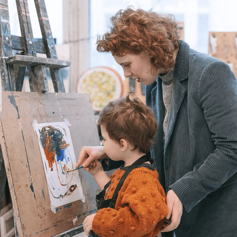 Woman helping young child paint on an easel.
